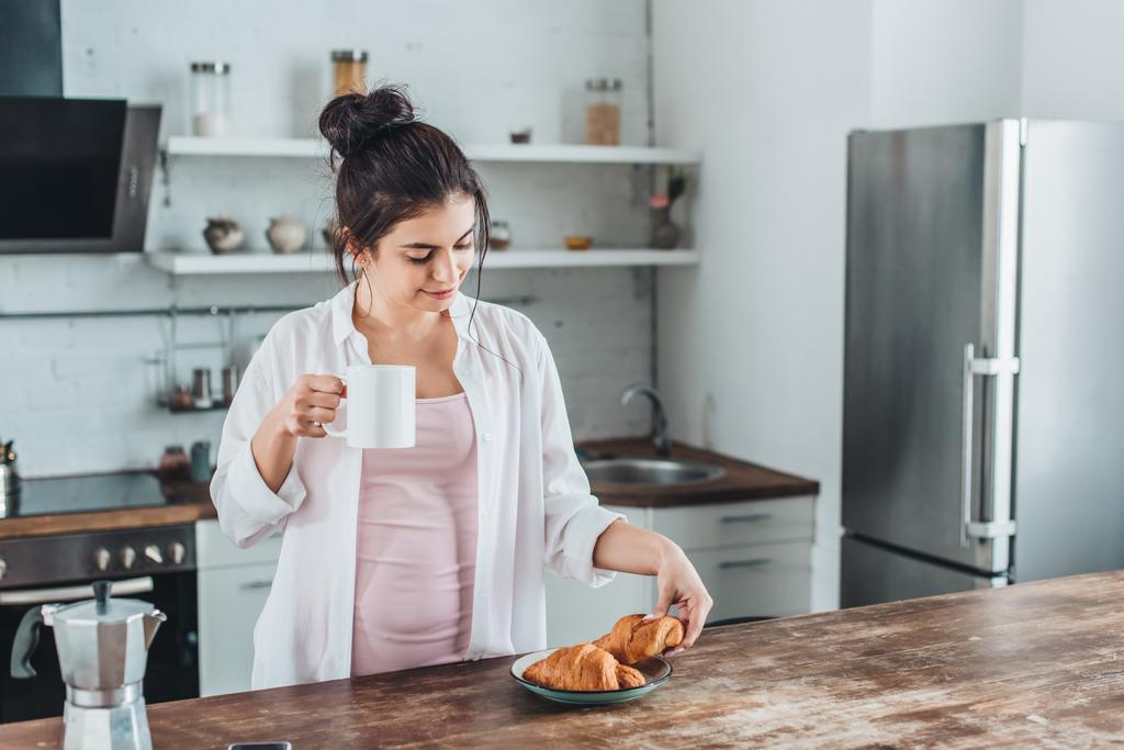 vrolijke jonge vrouw ontbijten met croissants en koffie aan houten tafel in de keuken thuis - Foto, afbeelding
