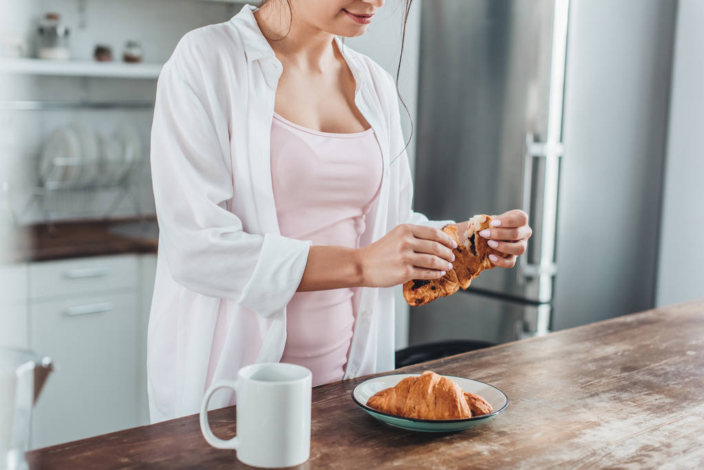 частичный вид женщины, завтракающей с круассанами и кофе за деревянным столом на кухне дома
 - Фото, изображение