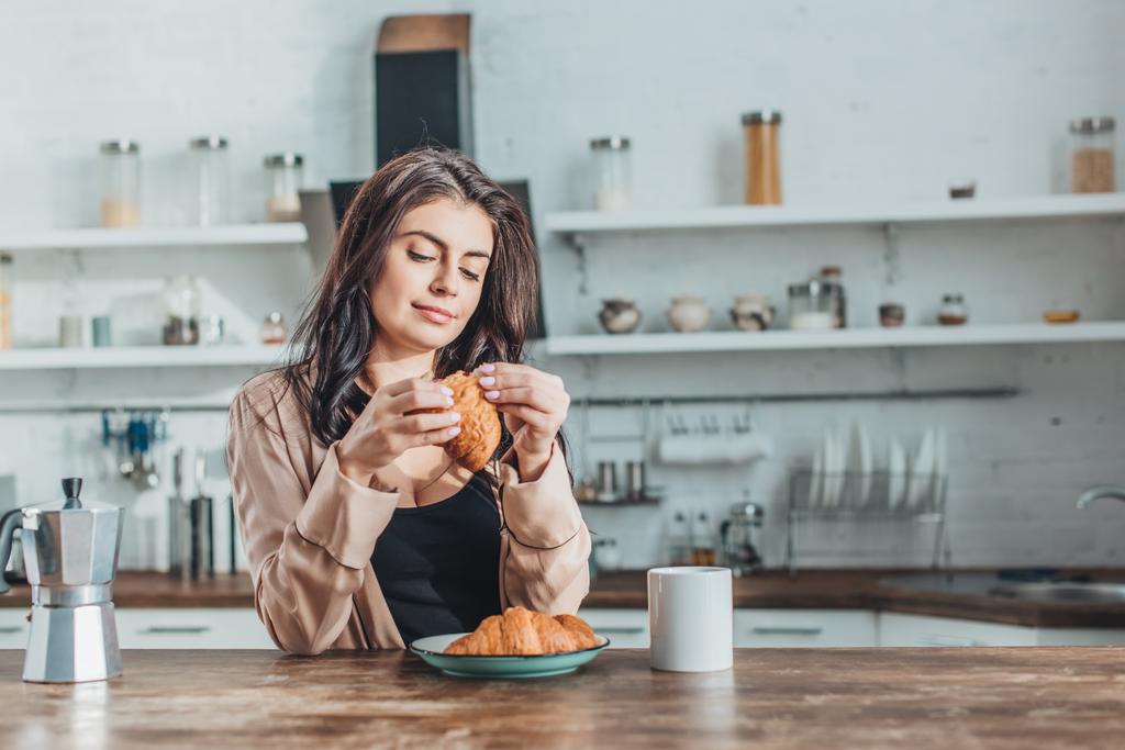 自宅の台所で木製のテーブルでクロワッサンとコーヒーの朝食を持つ若い美しい女性 - 写真・画像