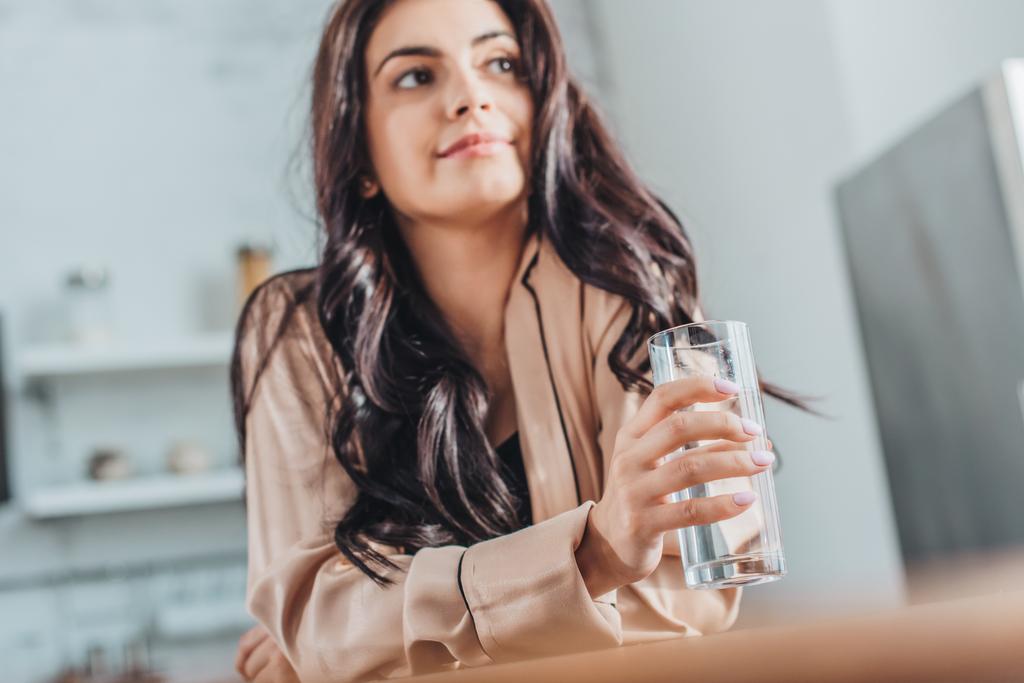 nízký úhel pohled krásná mladá žena drží sklenici s vodou a sedí na dřevěnou desku v kuchyni doma - Fotografie, Obrázek