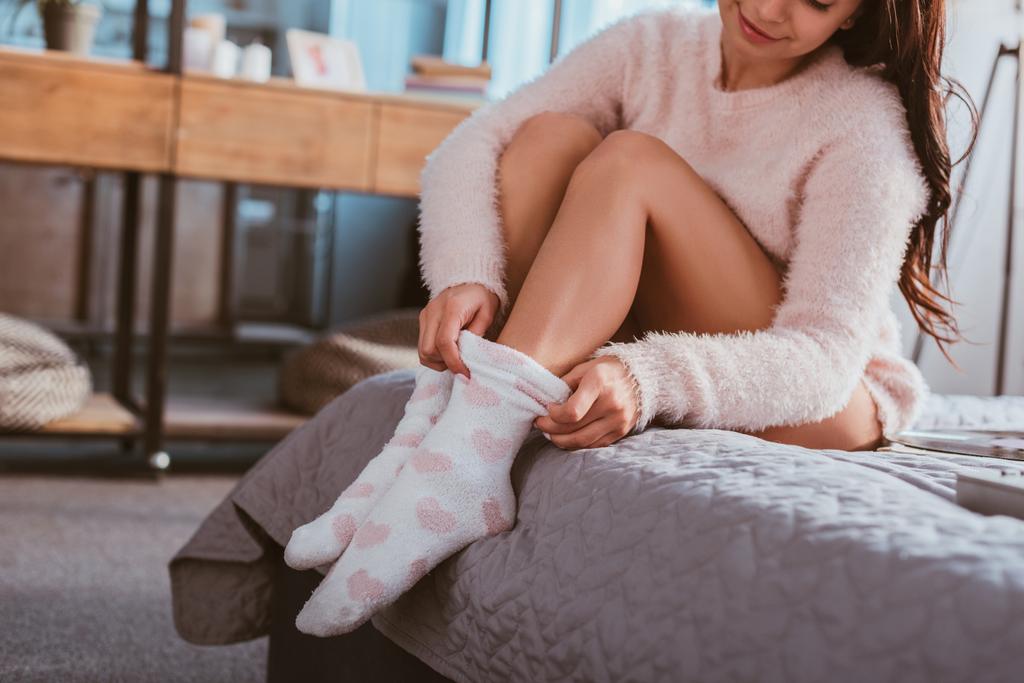 частичный вид девушки, надевающей пушистые носки, сидящей дома на кровати
 - Фото, изображение