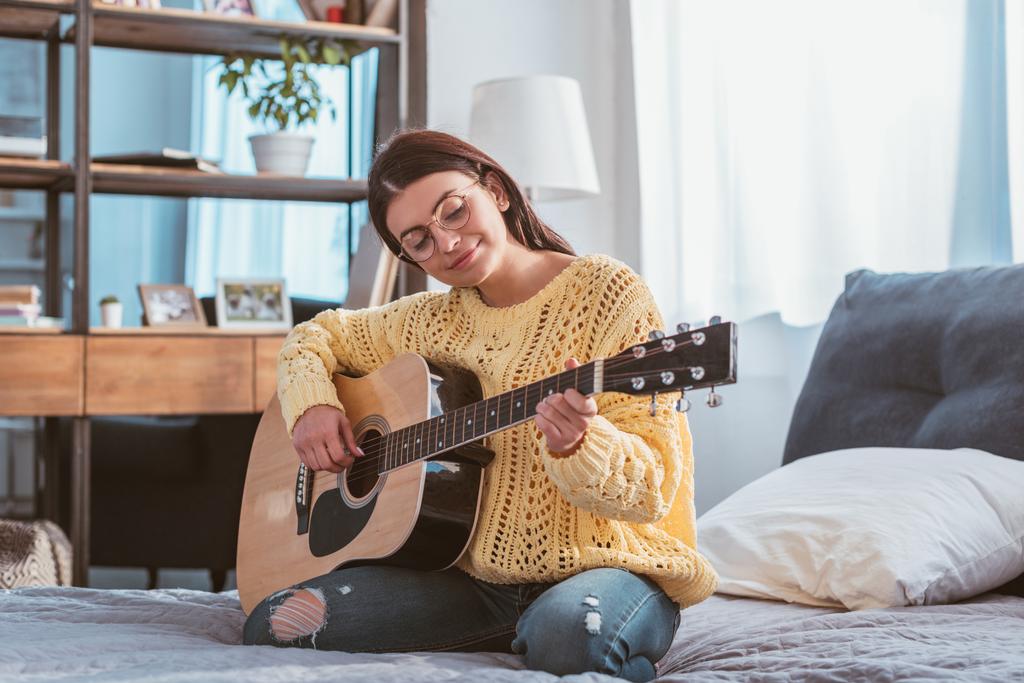 Улыбающаяся красивая женщина в очках, играющая на акустической гитаре, сидя дома на кровати
 - Фото, изображение