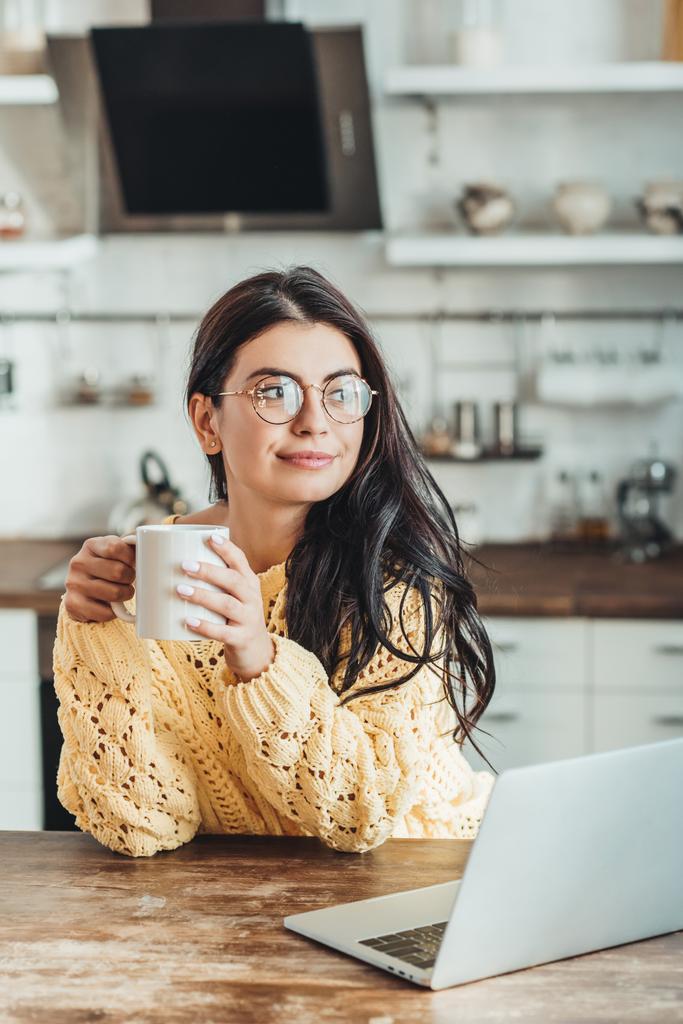 vrolijke vrouwelijke freelancer in brillen met koffiekopje zittend aan houten tafel met laptop in de keuken thuis - Foto, afbeelding