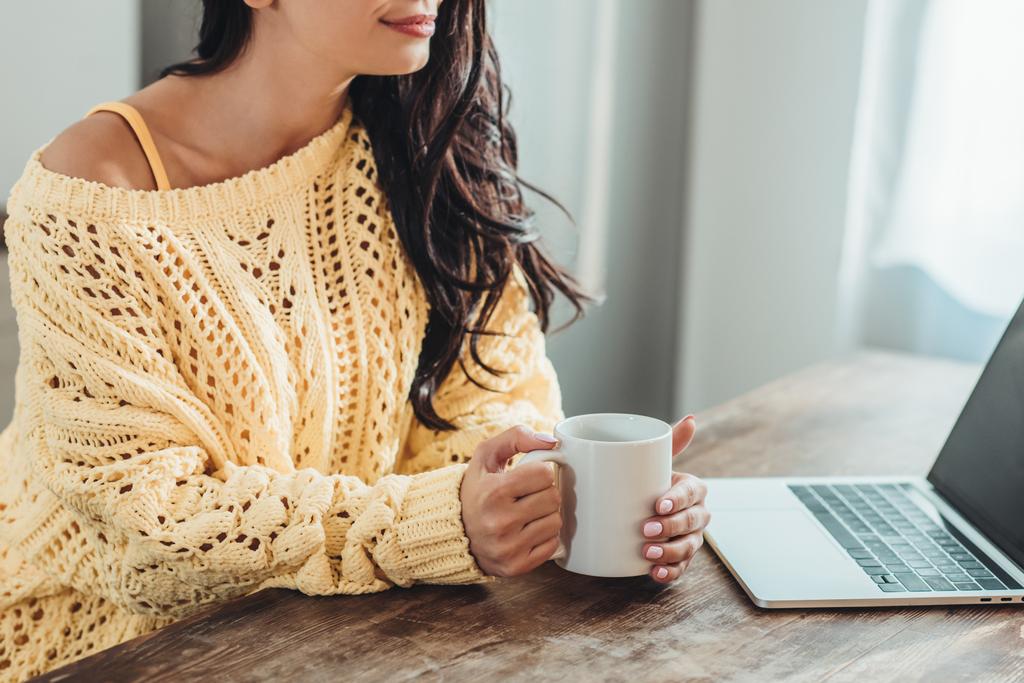 vista parziale di freelance donna in possesso di una tazza di caffè e seduta a tavolo in legno con computer portatile in cucina a casa
 - Foto, immagini
