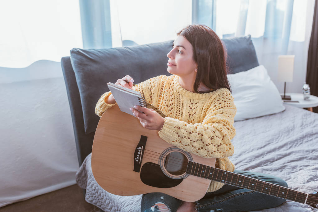 Szczęśliwa dziewczyna siedzi z gitara akustyczna i podręcznik na łóżko w domu - Zdjęcie, obraz