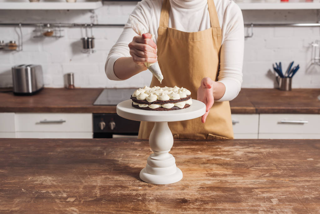 bijgesneden schot van vrouw in schort versieren gastronomische zoet gebak met slagroom    - Foto, afbeelding