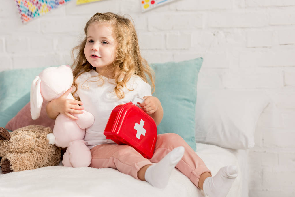 adorable kid holding first aid kit and rabbit toy in children room, looking away - Photo, Image
