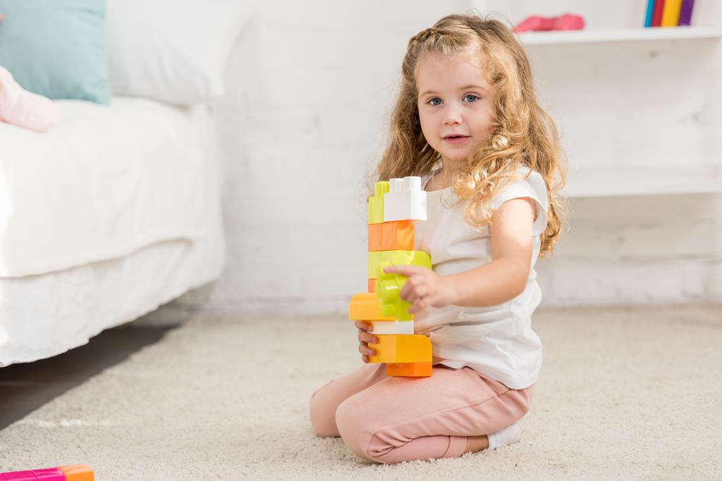 чарівна дитина грає з кольоровим пластиковим конструктором на килимі в дитячій кімнаті і дивиться на камеру
 - Фото, зображення