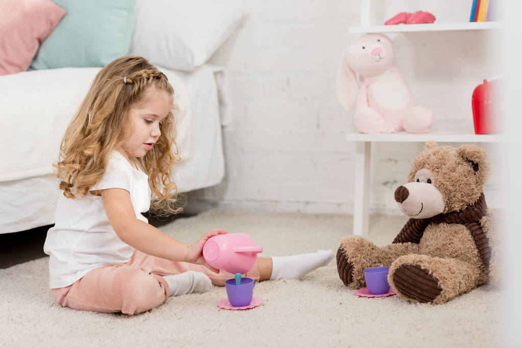 αξιολάτρευτο παιδί παίζει με αρκουδάκι και πλαστικά ποτήρια στο πάτωμα στο δωμάτιο παιδιών - Φωτογραφία, εικόνα