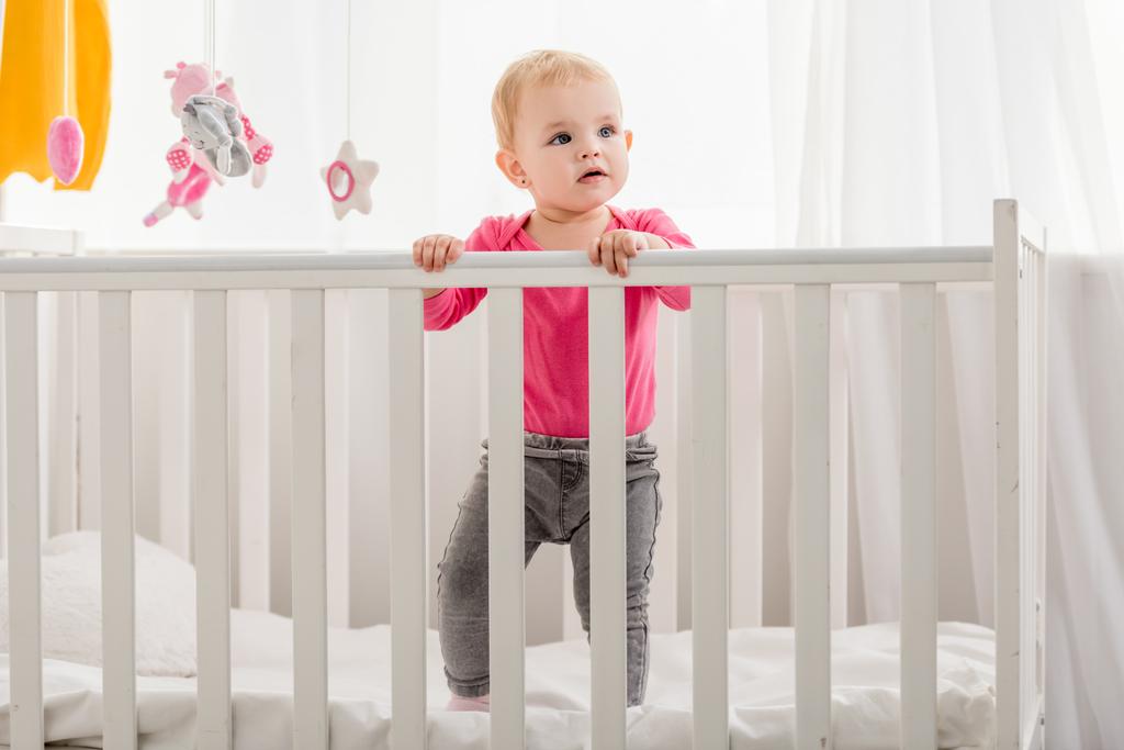 adorable kid in pink shirt standing in crib and looking away - Photo, Image