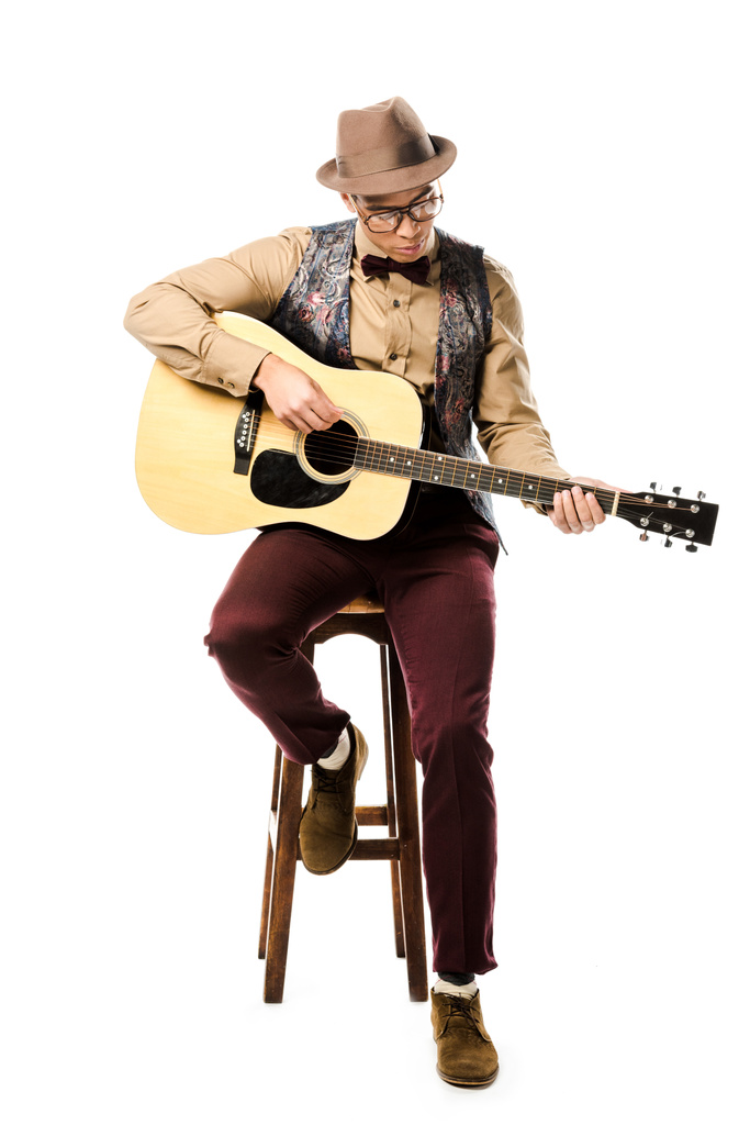 músico masculino de raza mixta enfocado en sombrero y anteojos tocando en guitarra acústica mientras está sentado en silla aislado en blanco
 - Foto, Imagen
