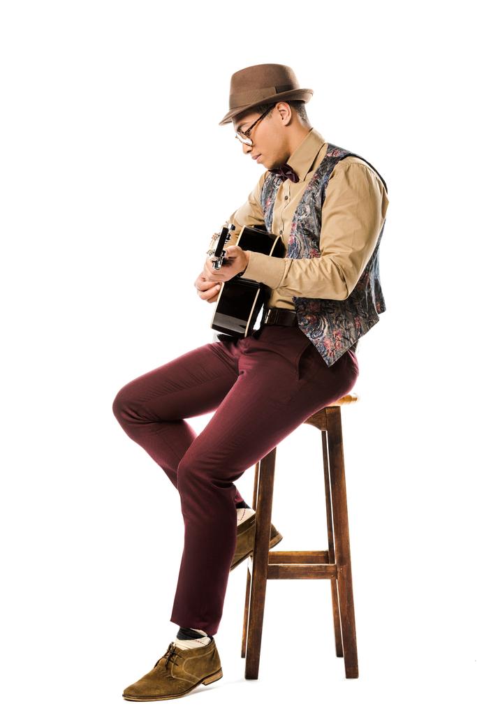side view of mixed race male musician in hat and eyeglasses playing on acoustic guitar while sitting on chair isolated on white - Photo, Image