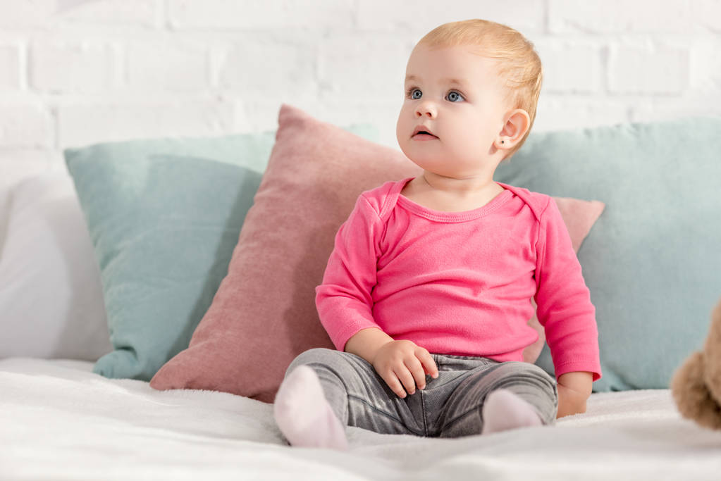 entzückendes Kind, das mit Kissen auf dem Bett sitzt und im Kinderzimmer aufblickt - Foto, Bild