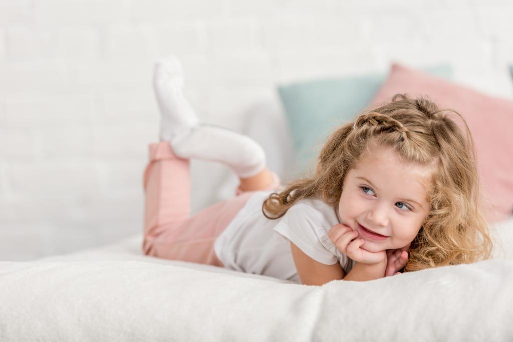 alegre adorable feliz niño acostado en la cama en la habitación de los niños y mirando hacia otro lado
 - Foto, Imagen