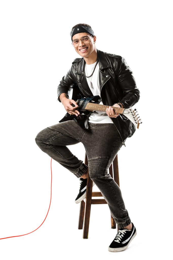 γελώντας αρσενικό μουσικός σε δερμάτινο μπουφάν που παίζει στην ηλεκτρική κιθάρα, ενώ κάθεται στην καρέκλα που απομονώνονται σε λευκό - Φωτογραφία, εικόνα