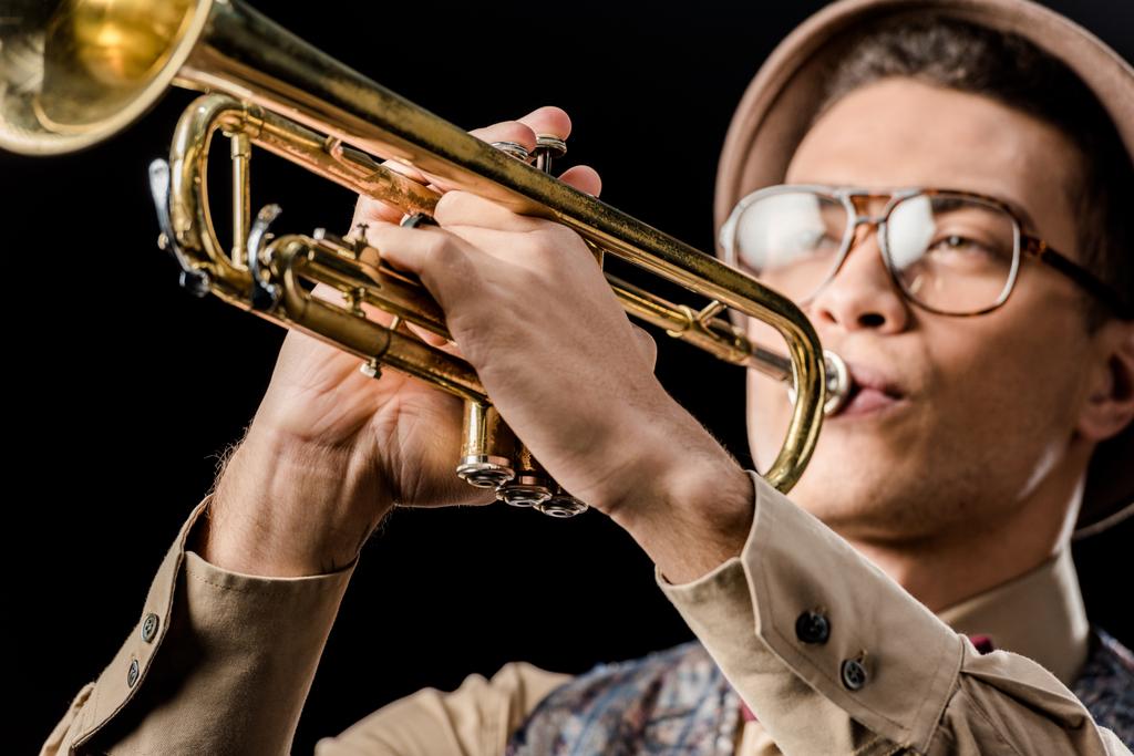 close up view of mixed race male jazzman in hat and eyeglasses playing on trumpet isolated on black - Photo, Image
