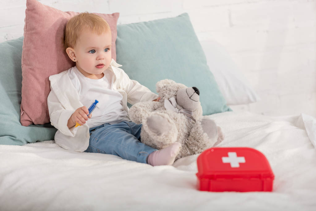 entzückendes Kind sitzt mit Teddybär und Verbandskasten im Kinderzimmer auf dem Bett - Foto, Bild