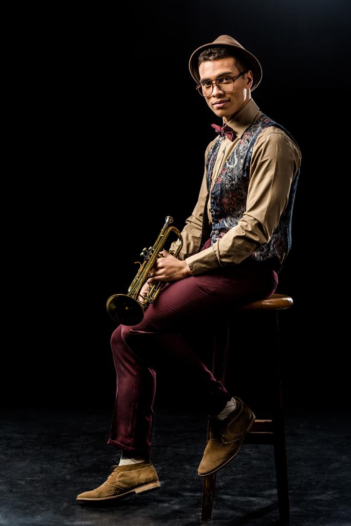 sonriente mestizo masculino de raza mixta posando con trompeta mientras está sentado en silla en negro
  - Foto, Imagen