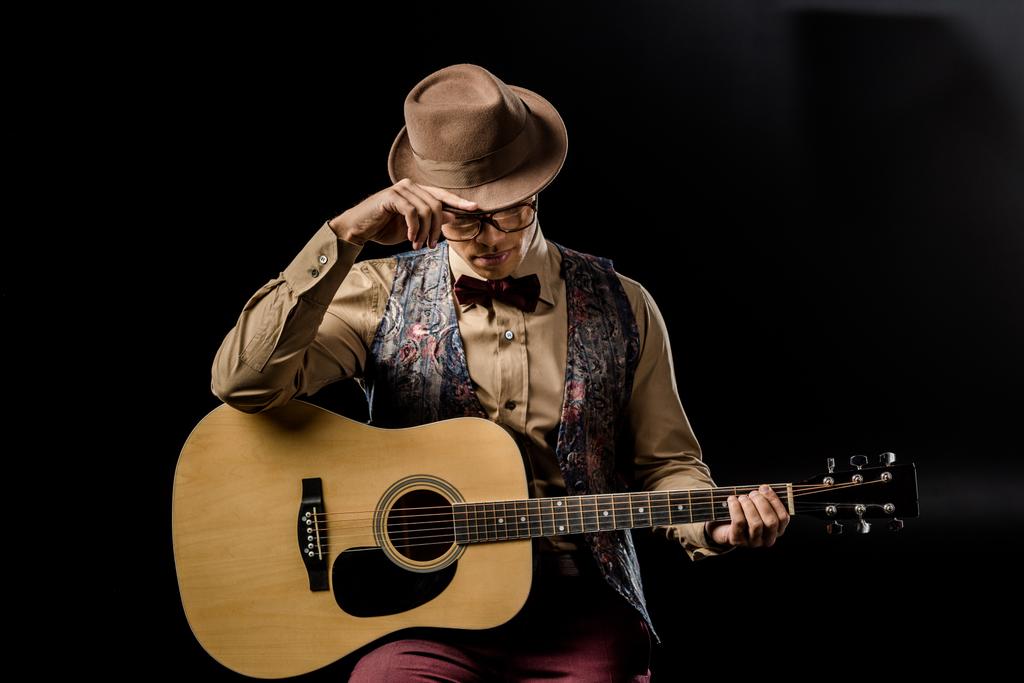 stylische gemischte Rasse männliche Musiker mit Brille und Hut posiert mit Akustikgitarre isoliert auf schwarz - Foto, Bild