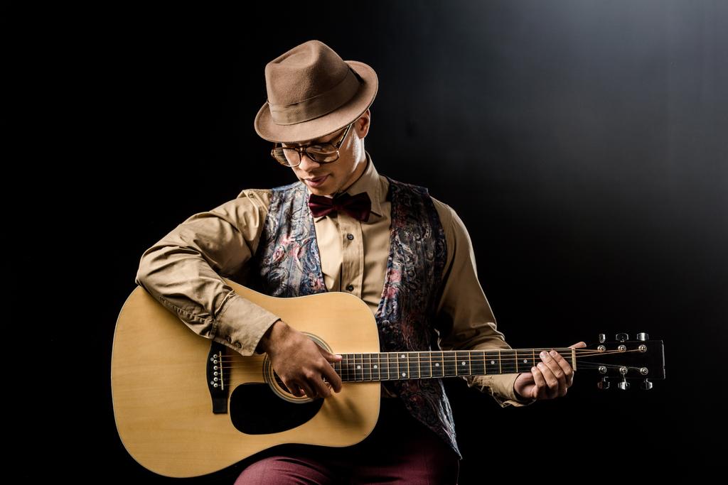 Молодой красивый мужчина в очках и шляпе, играющий на акустической гитаре, изолированной на черном
 - Фото, изображение