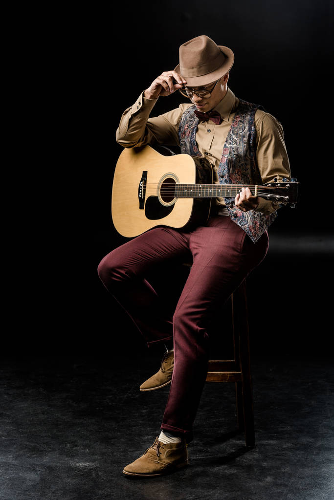 стильний молодий чоловік в капелюсі позує з акустичною гітарою, сидячи на стільці на чорному
  - Фото, зображення