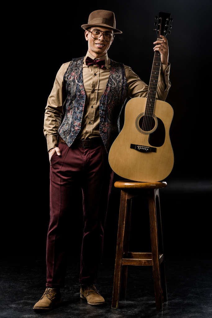 mužské hudebník v stylovém klobouku pózuje s akustickou kytarou poblíž židle na černém pozadí  - Fotografie, Obrázek