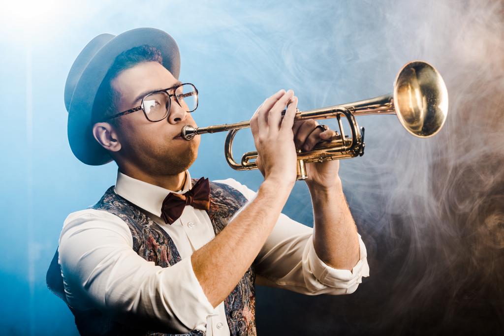 stilvoller Jazzer mit Hut und Brille, der auf der Bühne mit dramatischer Beleuchtung und Rauch auf der Trompete spielt - Foto, Bild