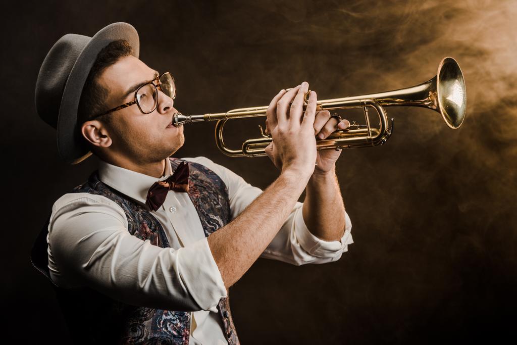 jong gemengd ras jazzman in hoed en bril op trompet spelen op het podium met dramatische verlichting en rook - Foto, afbeelding