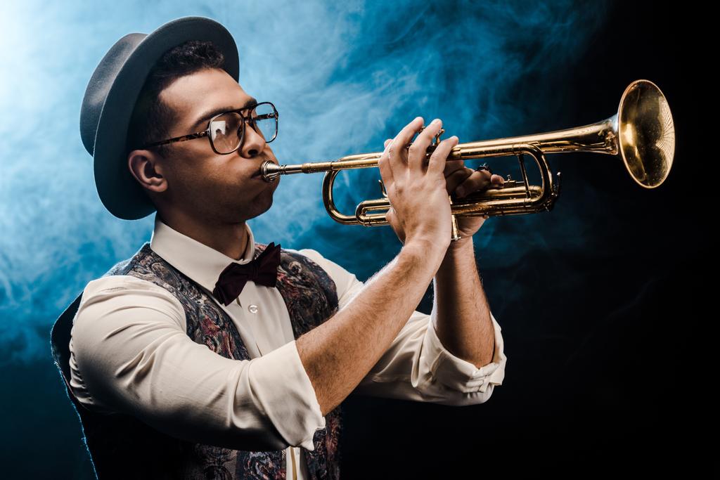 mannelijke zanger in hoed en bril op trompet spelen op het podium met dramatische verlichting en rook - Foto, afbeelding