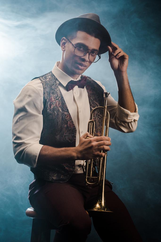 şapka ve gözlük duman ve dramatik aydınlatma ile sahnede trompet ile poz yakışıklı şık genç adam  - Fotoğraf, Görsel