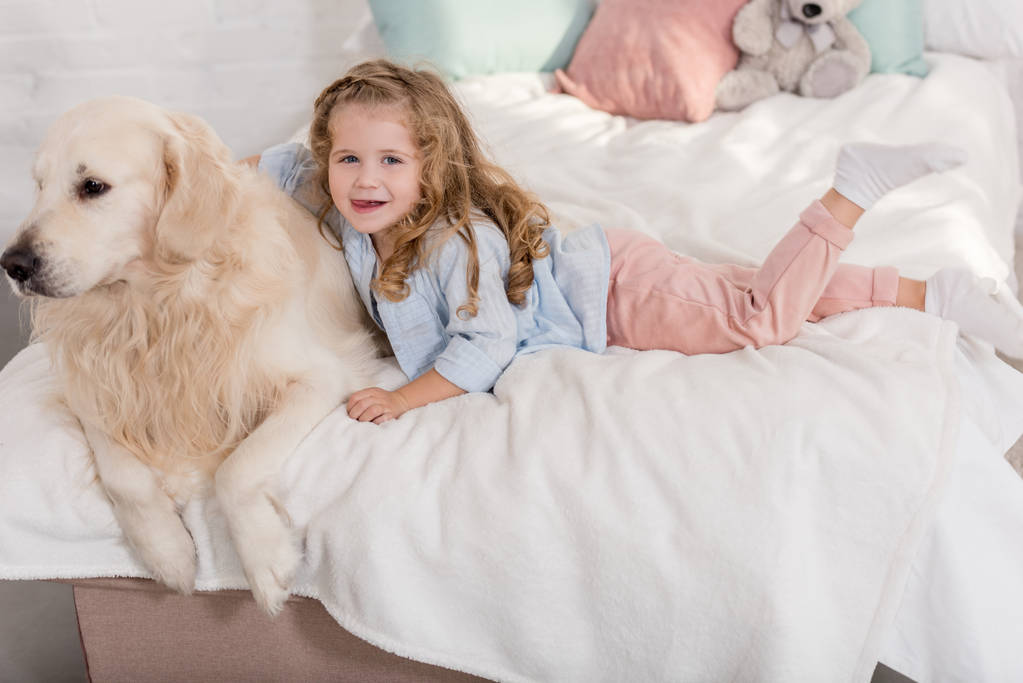 wysoki kąt widzenia adorable kid przytulanie golden retriever na łóżku w pokoju dzieci i patrząc na kamery - Zdjęcie, obraz
