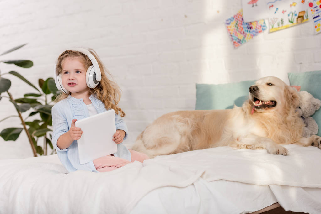 adorable niño escuchando música con la tableta, golden retriever acostado en la cama en la habitación de los niños
 - Foto, imagen