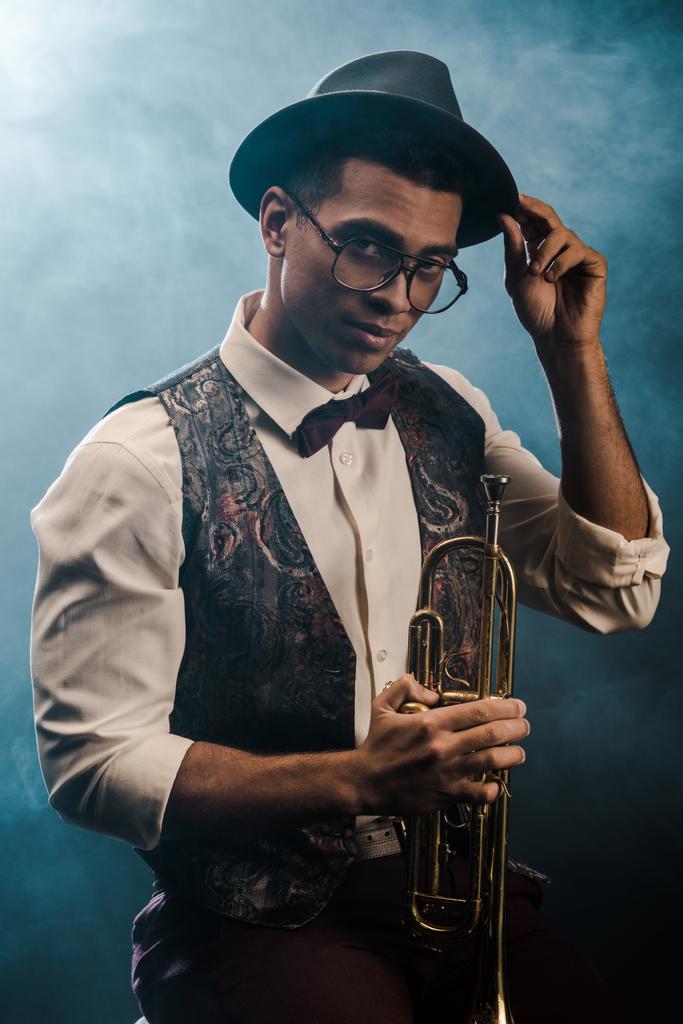 jeune homme élégant et confiant dans le chapeau et les lunettes posant avec trompette sur scène avec fumée et éclairage dramatique
  - Photo, image
