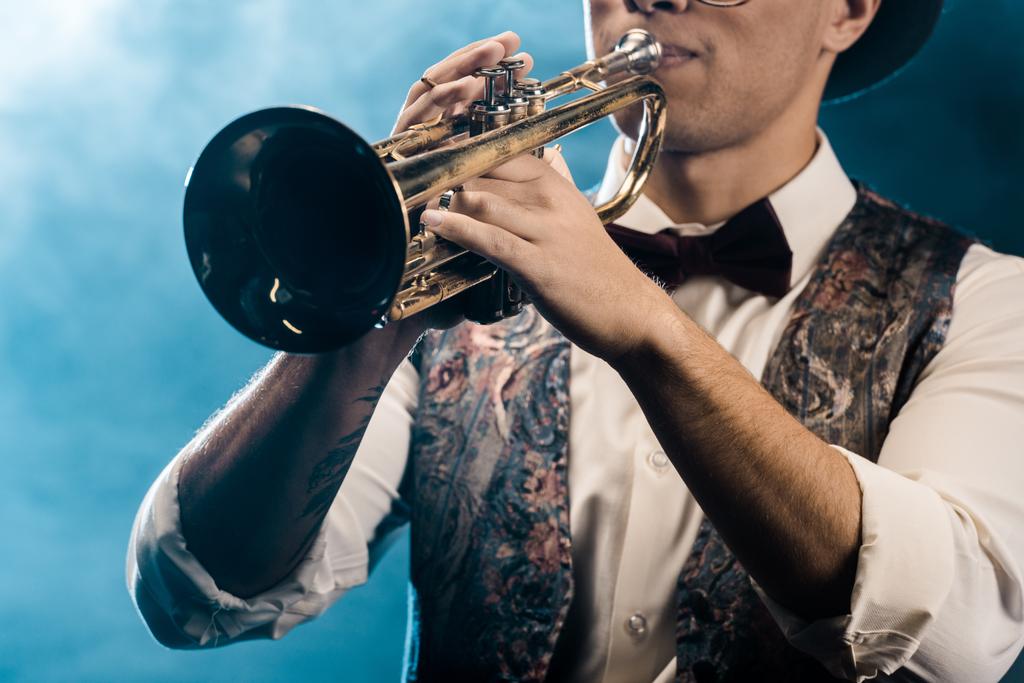 частичный вид мужчины-музыканта, играющего на трубе на сцене с драматическим освещением и дымом
 - Фото, изображение