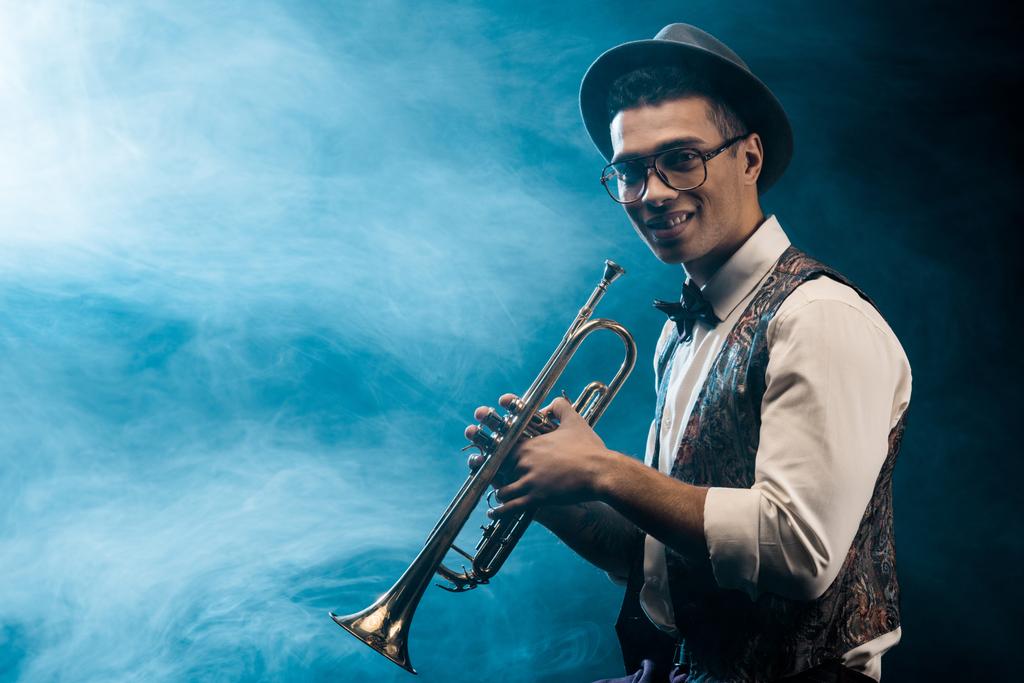 χαμογελώντας αρσενικό μουσικός ποζάρει με τρομπέτα στη σκηνή με δραματικό φωτισμό και τον καπνό - Φωτογραφία, εικόνα