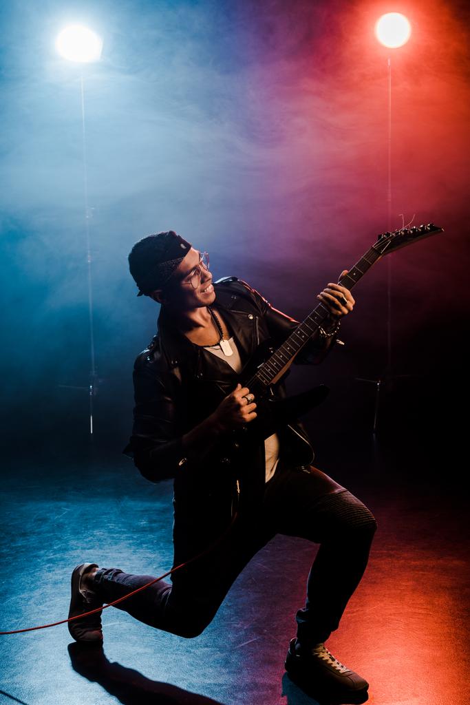 usmívající se muž rocková hvězda v kožené bundě plnění na elektrickou kytaru na jevišti s kouřem a dramatické osvětlení  - Fotografie, Obrázek