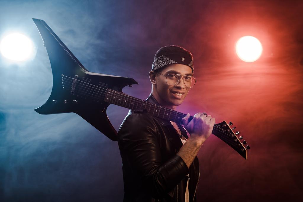smiling mixed race rock musician in leather jacket posing with electric guitar on stage with smoke and dramatic lighting - Photo, Image
