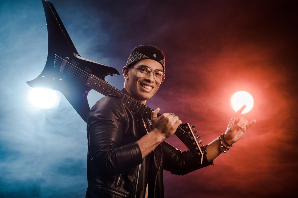 αρσενικό rocker σε δερμάτινο μπουφάν που παρουσιάζουν με ηλεκτρική κιθάρα και κάνει σημάδι κέρατα επί σκηνής με τον καπνό και δραματικό φωτισμό - Φωτογραφία, εικόνα