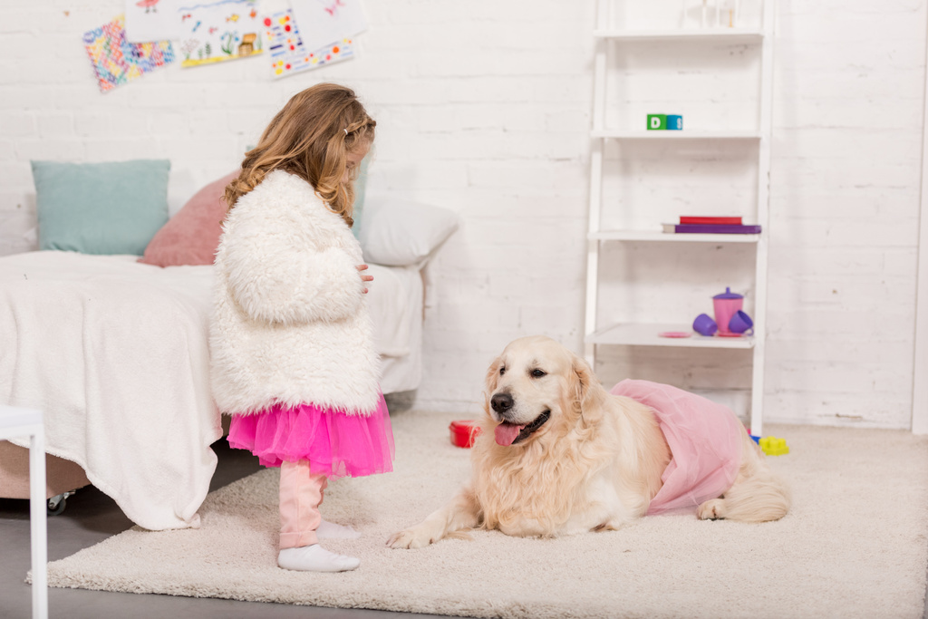 Kid in rok kijken naar golden retriever liggend op tapijt in roze rok in de kinderkamer - Foto, afbeelding
