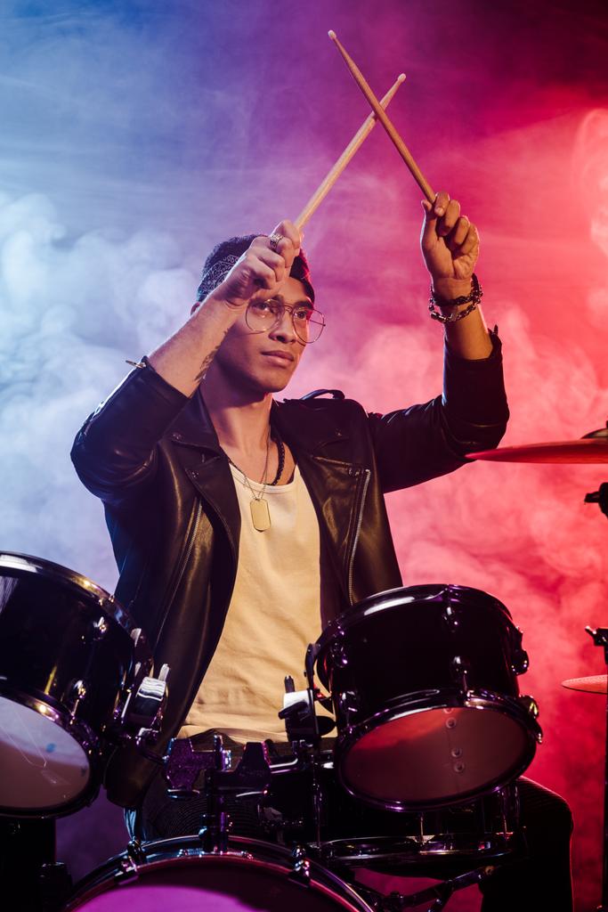 mixed race male musician raising drum sticks while sitting behind drum set on stage with dramatic lighting and smoke - Photo, Image