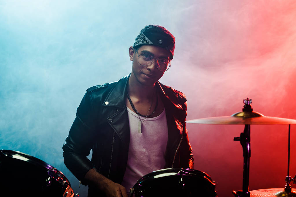 hezký mužský hudebník v kožené bundě sedí za bicí soupravu na jevišti s kouřem a dramatické osvětlení - Fotografie, Obrázek
