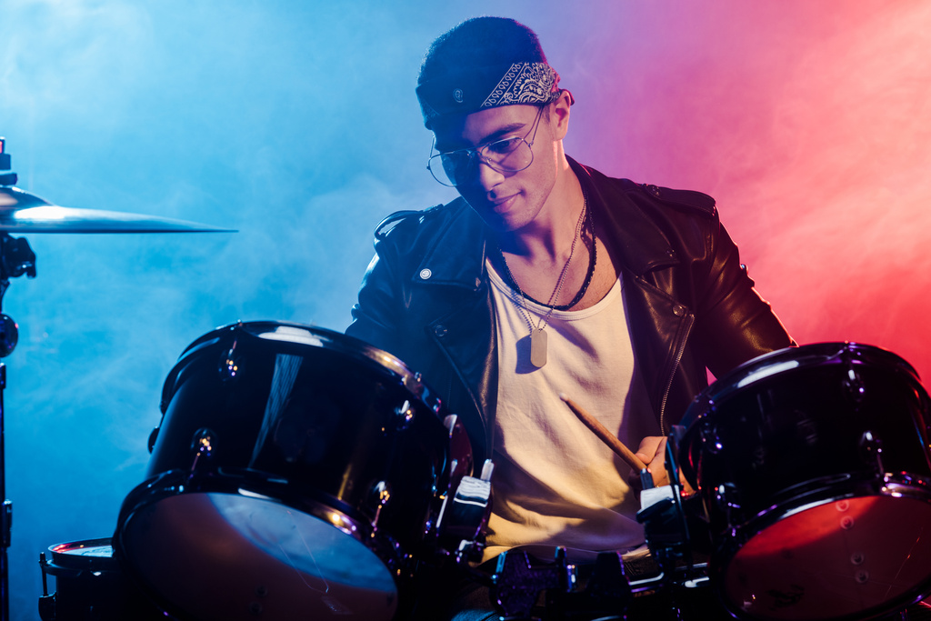 jovem de raça mista tocando bateria durante concerto de rock no palco com fumaça e iluminação dramática
 - Foto, Imagem