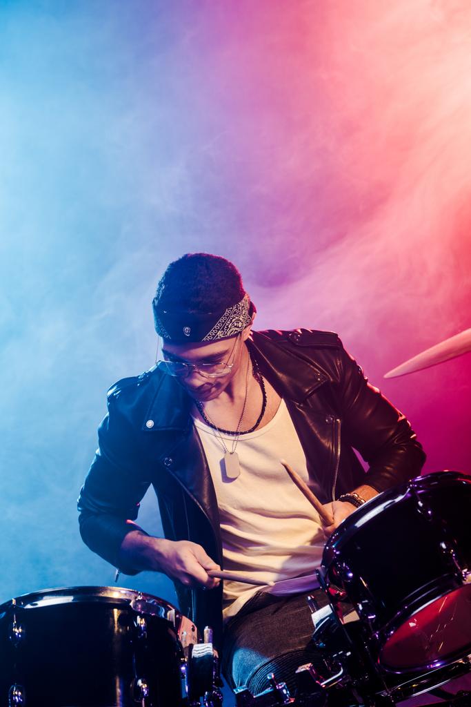 συγκεντρωμένη νεαρό αρσενικό μουσικός σε δερμάτινο μπουφάν παίζει ντραμς κατά τη διάρκεια συναυλίας ροκ στη σκηνή με τον καπνό και δραματικό φωτισμό - Φωτογραφία, εικόνα