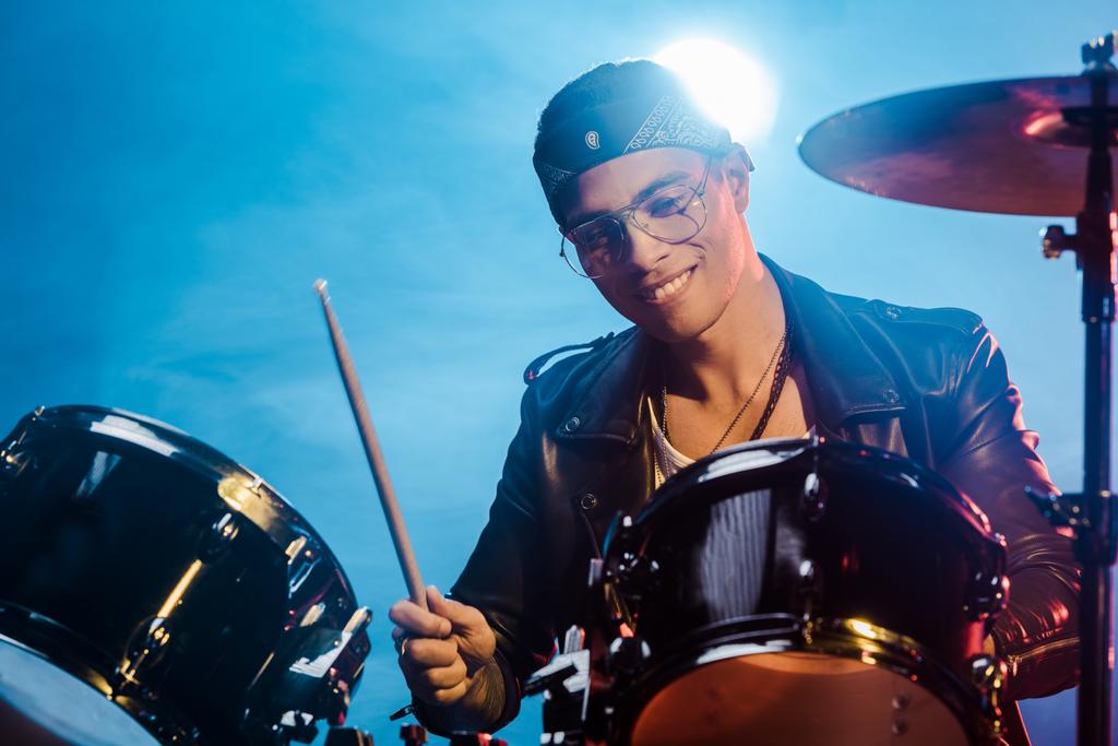 muzyk mężczyzna przystojny mieszanej rasy w skórzanej kurtce gry perkusji podczas koncertu rock na scenie z dymu i spotlight - Zdjęcie, obraz