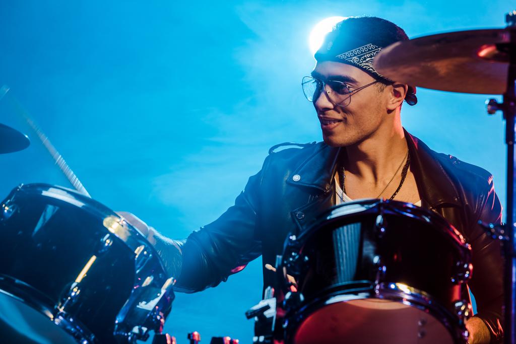 веселый мужчина-музыкант в кожаной куртке играет на барабанах во время рок-концерта на сцене с дымом и прожектором
 - Фото, изображение