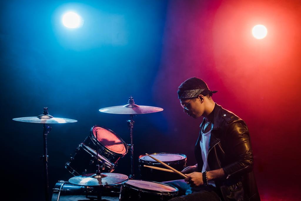мужчина-музыкант в кожаной куртке играет на барабанах во время рок-концерта на сцене с дымом и прожекторами
 - Фото, изображение