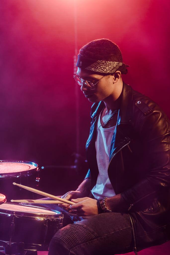 músico masculino em jaqueta de couro tocando bateria durante concerto de rock no palco com fumaça e iluminação dramática
  - Foto, Imagem