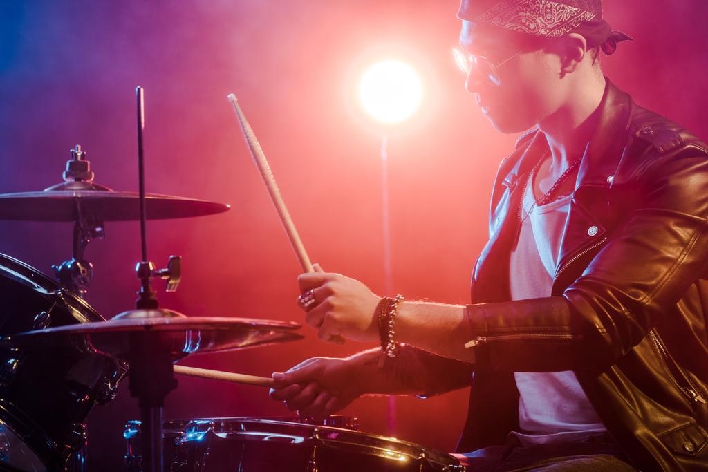 Widok z boku z młody muzyk mężczyzna w skórzanej kurtce gry perkusji podczas koncertu rock na scenie   - Zdjęcie, obraz