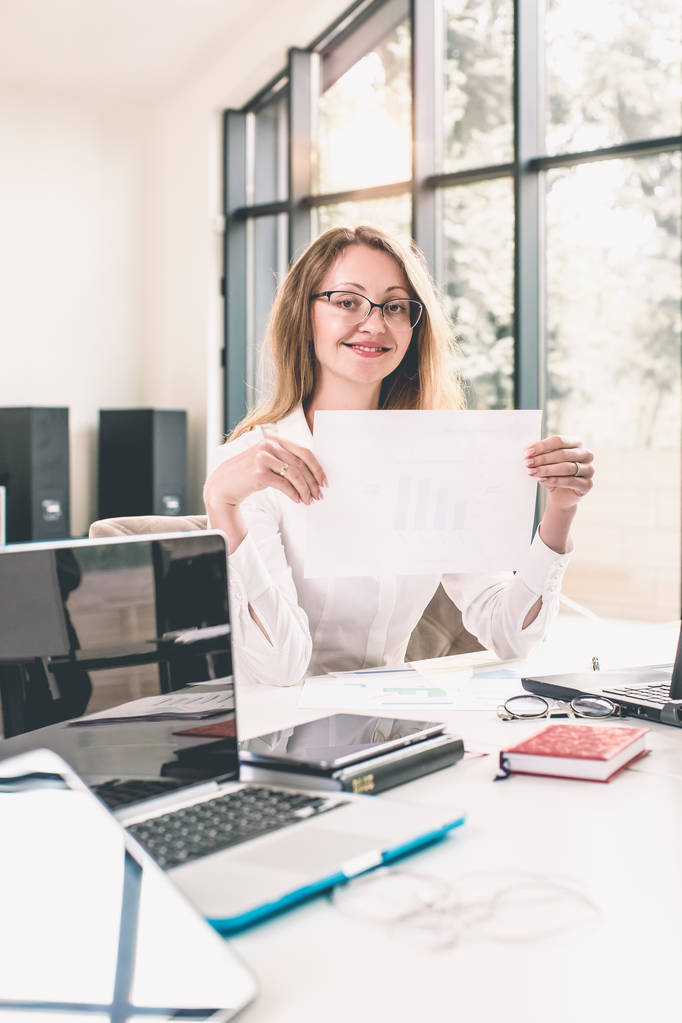 Succesvolle lachende volwassen zakenvrouw met behulp van laptop en computer terwijl het doen van wat papierwerk op kantoor - Foto, afbeelding