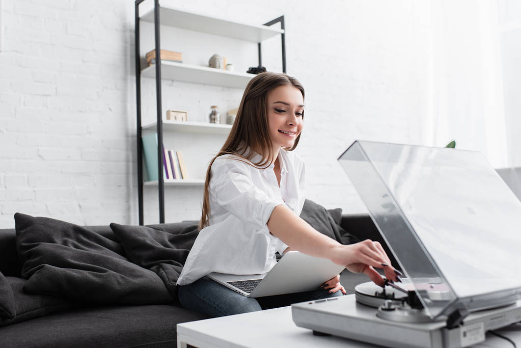 Lächelndes Mädchen in weißem Hemd sitzt auf Couch mit Laptop und spielt Schallplatte im Wohnzimmer ab - Foto, Bild